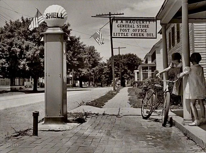 Black and white photo of two children with a bicycle outside W.R. Haggerty General Store and Post Office in Little Creek, Delaware. A vintage Shell gas pump and American flags are visible. Trees line the quiet street.