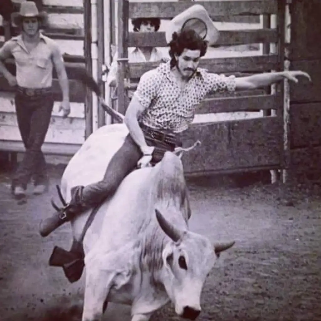 A rodeo rider in a cowboy hat and patterned shirt is riding a bucking bull. The rider balances with one arm up. The scene takes place in an outdoor arena with a person in the background watching. The image is in black and white.