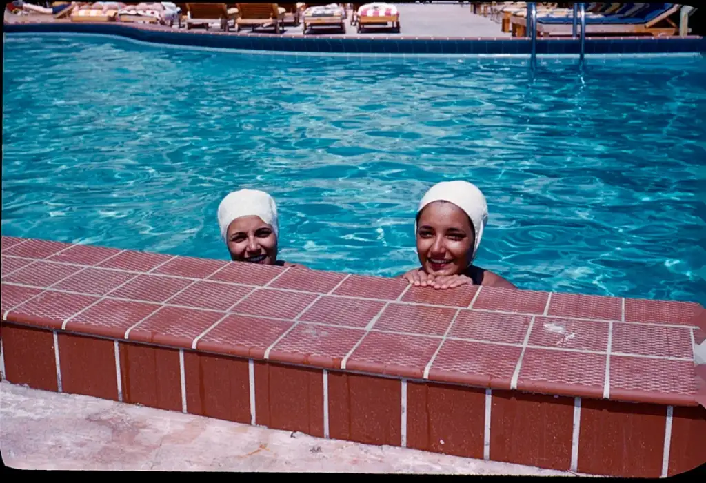 Two people wearing swimming caps are in a pool, smiling over the tiled edge. The water is clear, and loungers are visible in the background under a sunny sky.