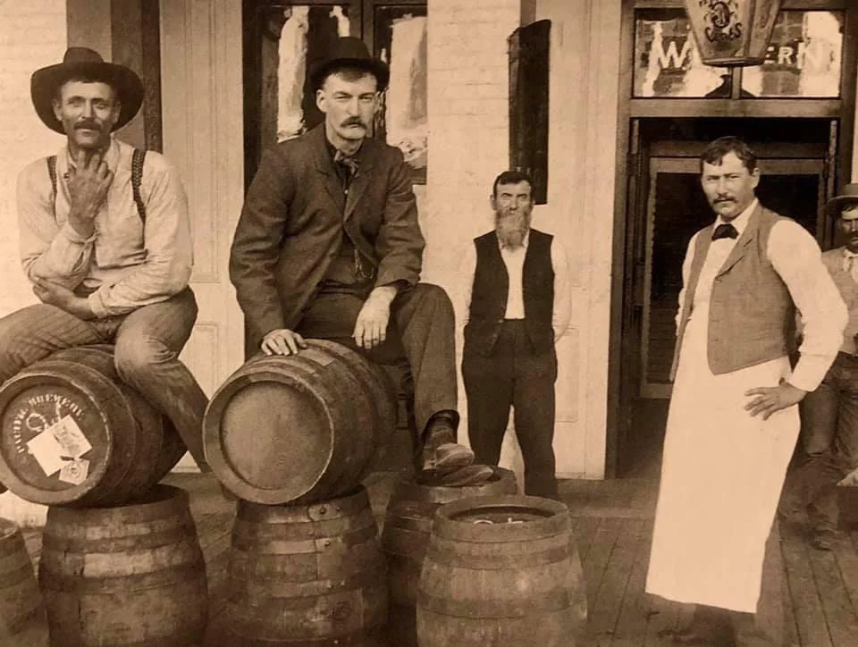 Four men pose with barrels in front of a building. Two sit on overturned barrels, one wears a hat and overalls, another in a suit. A bearded man stands in the background, while a man in a vest and apron stands to the side, hands on hips.