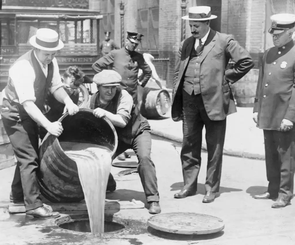 Men in 1920s attire empty a barrel of liquid onto the street, observed by three men in suits, one in a police uniform. A vintage vehicle and other men are in the background, evoking a Prohibition-era scene.