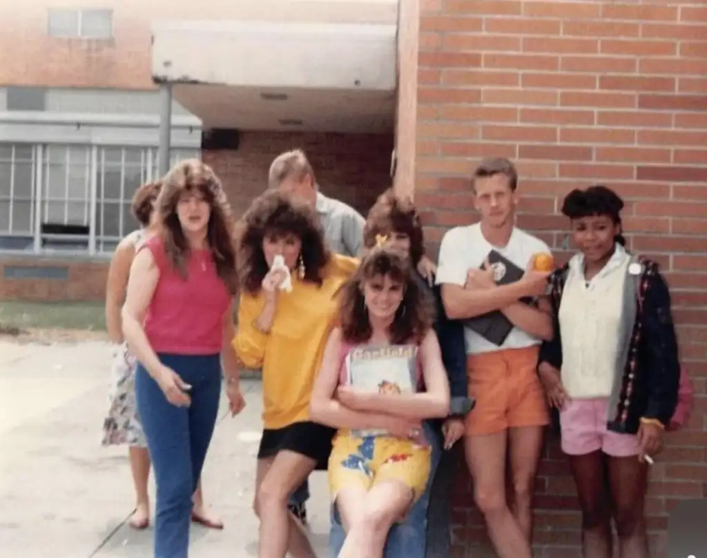 A group of seven people in 1980s fashion pose in front of a brick wall. Five women have big hairstyles, one in yellow is holding a drink. Two men are in shorts, one holds an orange. Outdoors, with a school window in the background.