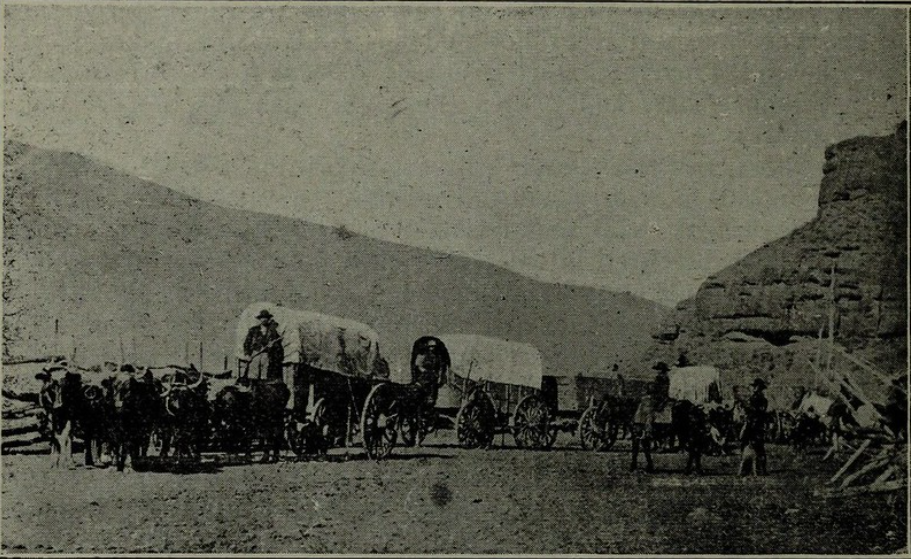 A vintage photo showing covered wagons lined up on a dirt path, drawn by horses or oxen. There are several figures, possibly cowboys, standing or sitting near the wagons. Rocky hills and a wooden fence are visible in the background.