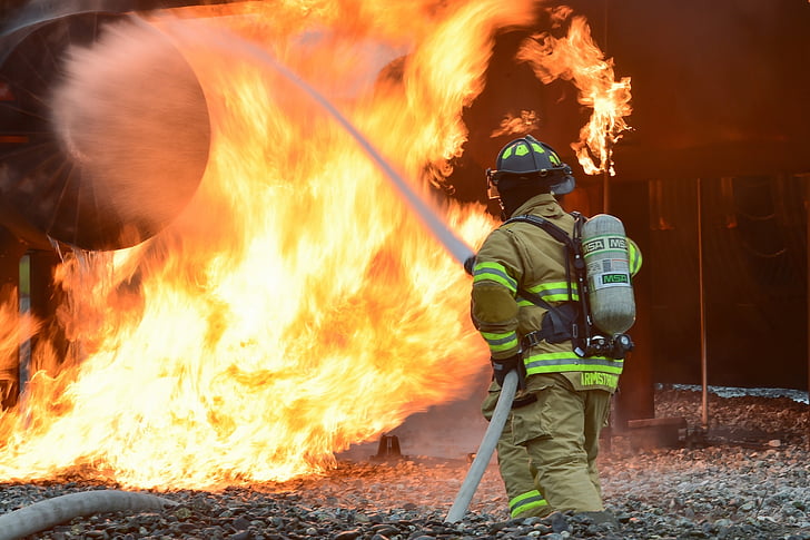 Firefighter in full gear sprays water from a hose at large, intense flames. The scene is outdoors, with the firefighter standing on rocky ground, battling the fire with determination.