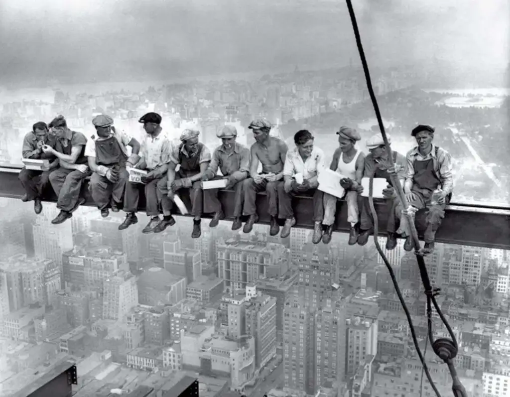 Black and white photo of eleven construction workers sitting on a steel beam high above a cityscape, eating lunch and chatting. They are suspended in the air with no safety harnesses, showcasing a foggy city below.