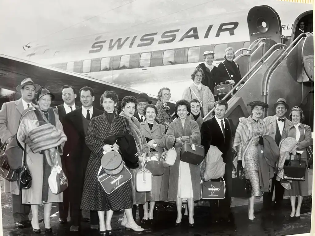 A group of well-dressed people, carrying bags and coats, stand in front of a Swissair airplane on the tarmac. Some are on the airplane stairs, while others pose at the bottom. The plane's name is visible in large letters.