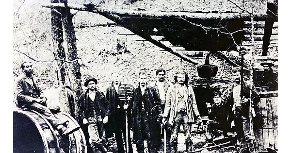 A black-and-white photo shows a group of men posing outdoors in front of industrial equipment, possibly related to mining or logging. They are dressed in period clothing, suggesting a historical setting.