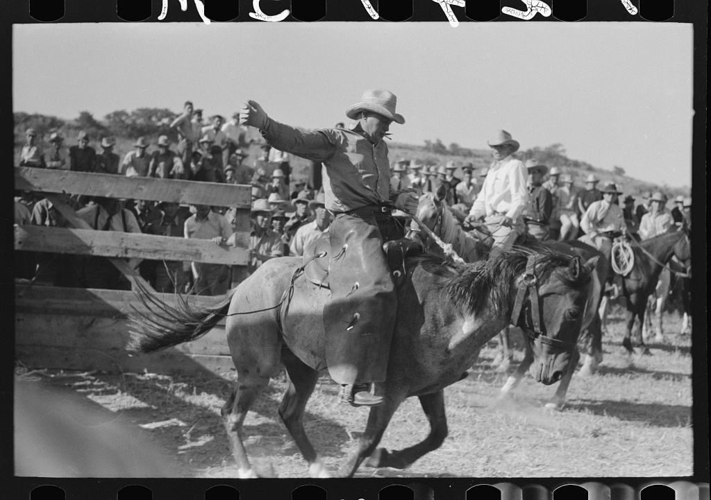 A cowboy rides a bucking horse during a rodeo event, with one arm raised for balance. The background features a crowd of spectators and wooden fencing under a clear sky. The rider wears a hat and traditional western attire.