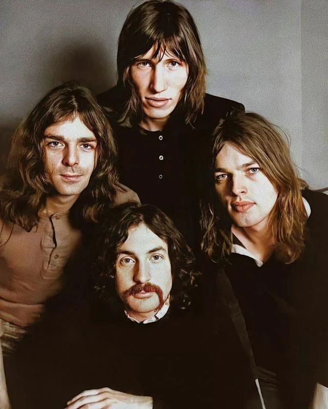 Four men with long hair pose together in a vintage-style portrait. The man at the front left has a mustache, while the others have clean-shaven faces. They are dressed in casual, dark clothing, set against a neutral background.