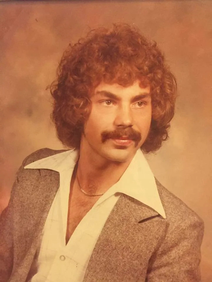 Portrait of a person with curly hair and a mustache, wearing a grey blazer over a white collared shirt. The background is a warm, brown gradient. The style is reminiscent of 1970s fashion.