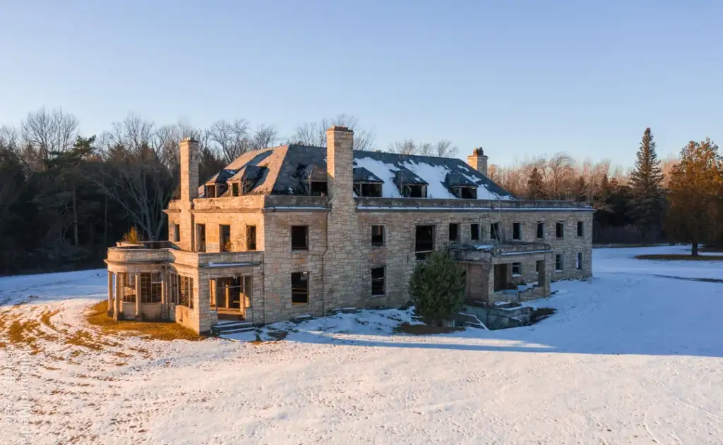 A large, abandoned stone building partially covered in snow. The structure has two prominent chimneys and numerous windows, some damaged. Surrounding it are snow-covered grounds with leafless trees in the background under a clear blue sky.