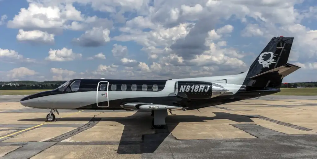 A sleek, black and silver private jet is parked on a runway under a partly cloudy sky. The registration number "N818RJ" is visible on its tail. The surroundings include distant trees and a clear horizon.