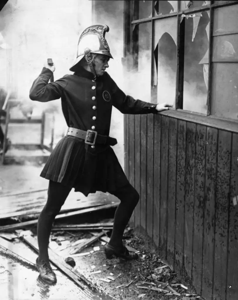 A vintage image of a firefighter in an old-fashioned uniform, including a helmet, coat, and skirt-like garment. The firefighter is using an axe to break a window, surrounded by debris.