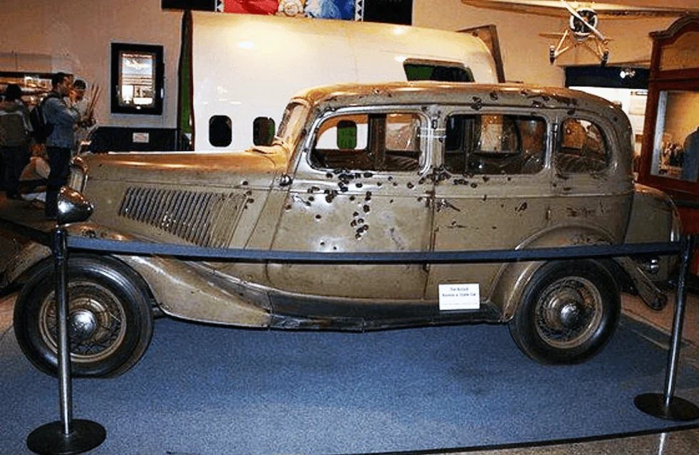 Brown vintage car riddled with bullet holes on display in a museum. The car is behind a black rope barrier, with a small information plaque in front. Other museum exhibits and people are visible in the background.