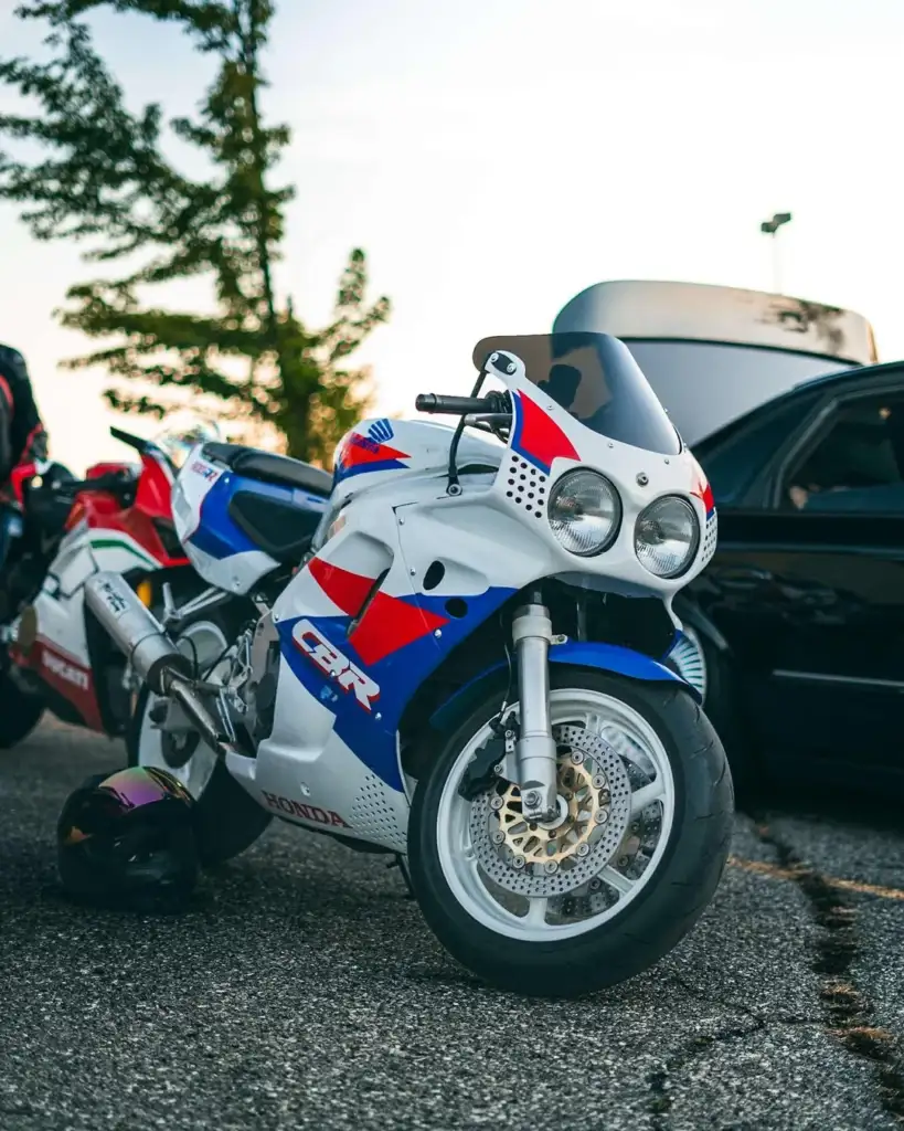 A vintage Honda CBR motorcycle in white with red and blue accents is parked on a paved surface near a black car. A helmet rests on the ground beside it. Trees and a reflective structure are visible in the background.