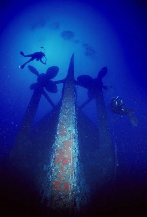 Divers explore the sunken hull of a ship, focusing on the large, twin propellers. The scene is illuminated by a deep, blue underwater light, adding to the mysterious atmosphere.