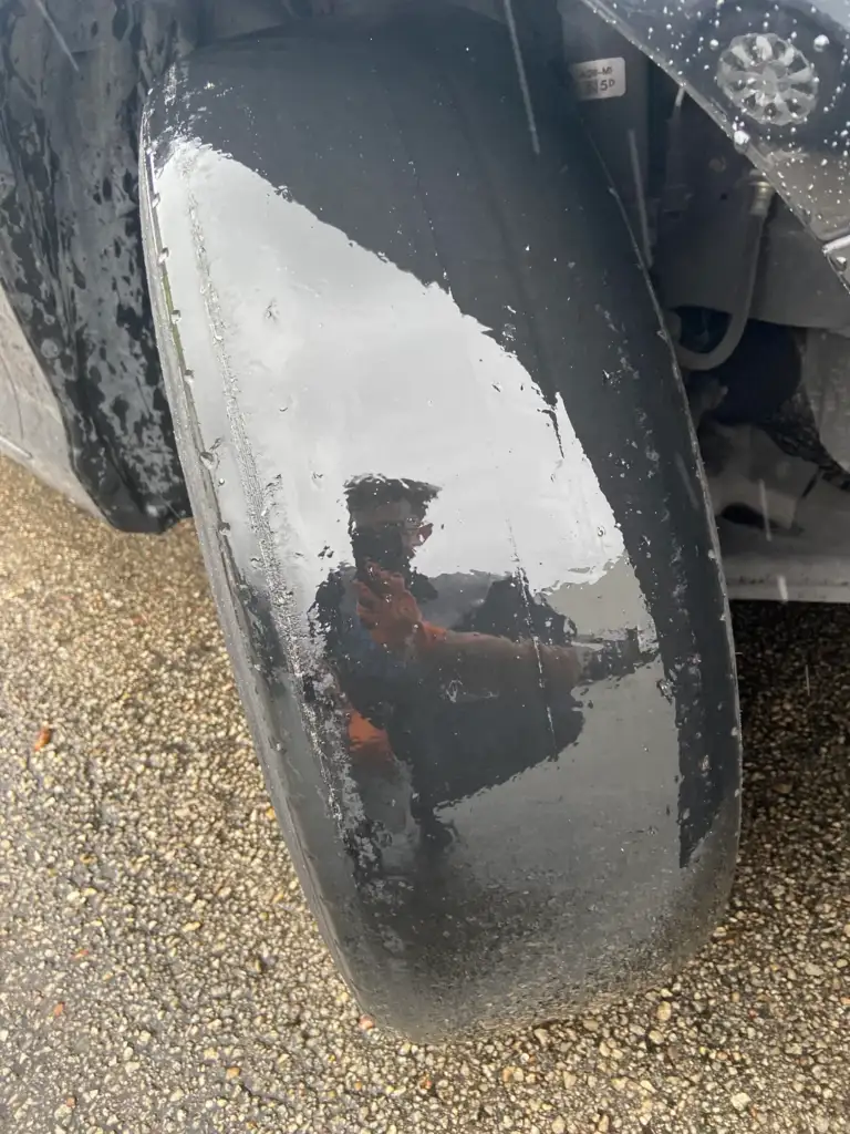 Close-up of a worn, smooth airplane tire with a reflective surface showing the faint reflection of a person wearing an orange vest. The tire is on a gravel surface, and droplets of water are visible on its surface.