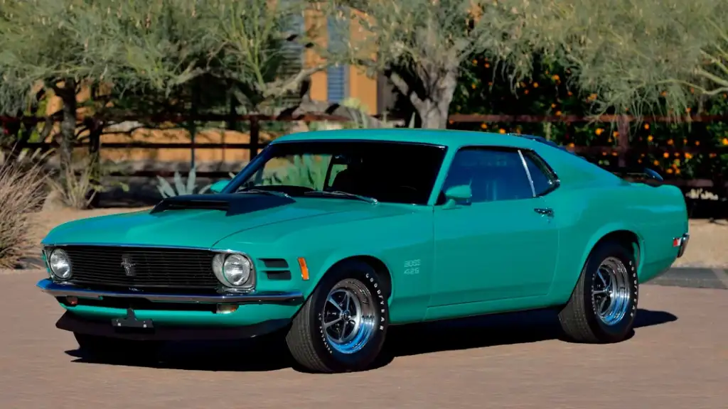 A teal classic muscle car, likely a 1970 Ford Mustang, is parked outdoors on a paved area. The car features a prominent hood scoop, chrome wheels, and is surrounded by desert vegetation.