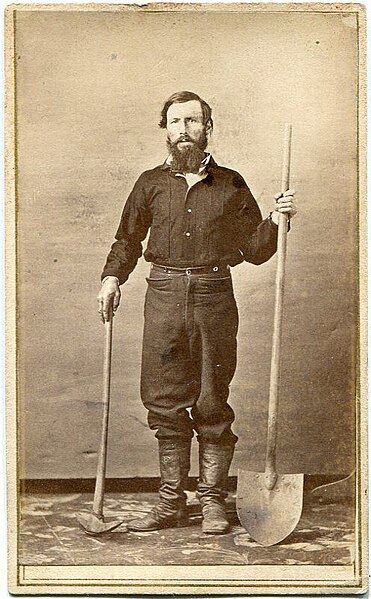 A sepia-toned vintage photograph of a man with a beard standing, holding a long-handled shovel and a pickaxe. He wears dark pants, a shirt, and boots, and poses against a simple backdrop.