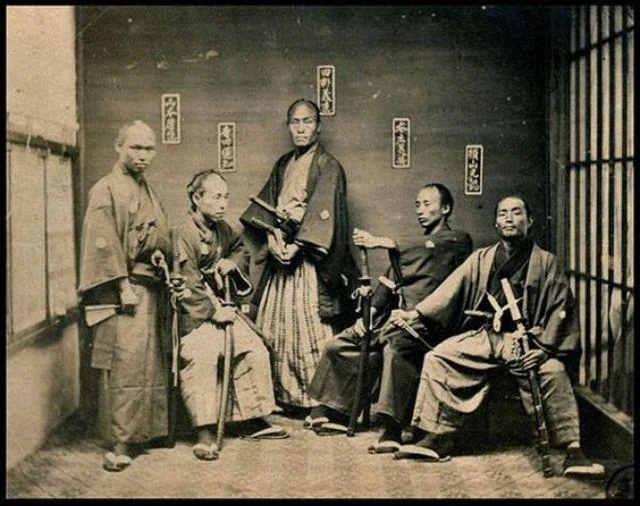 A vintage photograph of five samurai, dressed in traditional attire with swords. They are seated and standing in a room with wooden floors and walls, decorated with calligraphy. The image is in sepia tone, adding an antique feel.