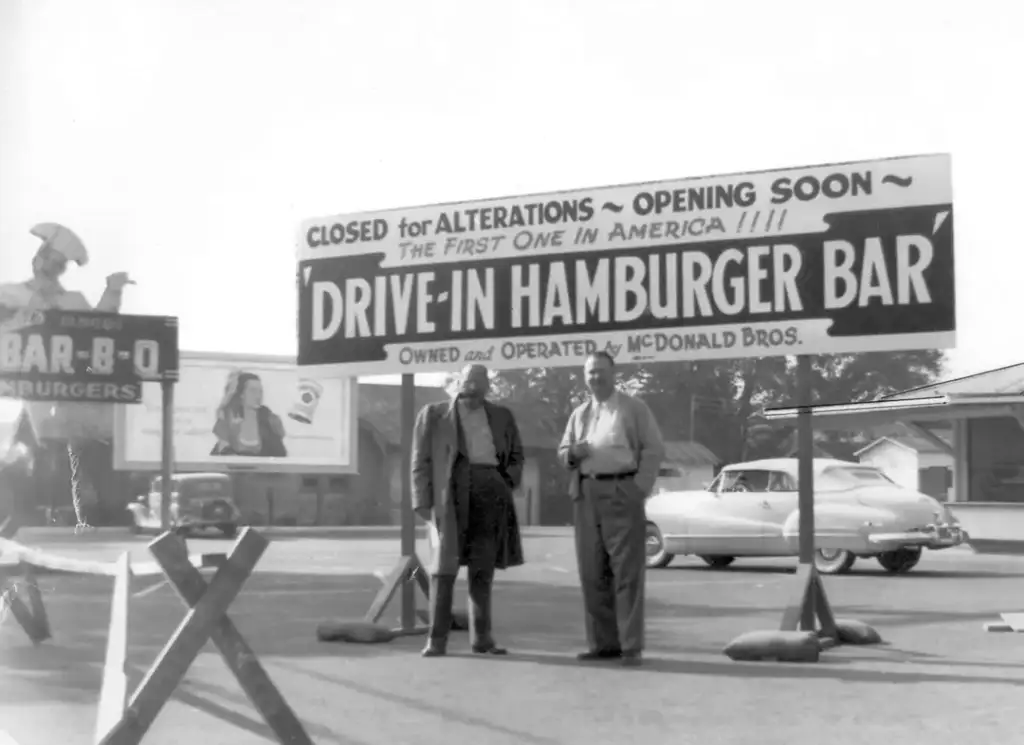 Two men stand in front of a large sign reading "Closed for Alterations ~ Opening Soon ~ 'Drive-In Hamburger Bar' ~ First One in America!!!" A vintage car and a billboard advertising BBQ hamburgers are visible in the background.