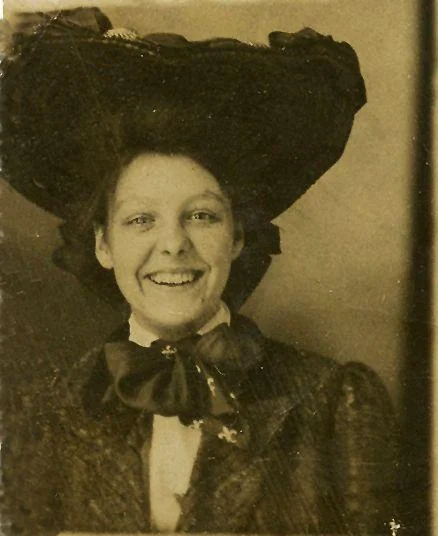 A sepia-toned vintage portrait of a woman smiling broadly. She wears a large, elaborate dark hat, a bow tie, and a textured jacket. The background is plain, highlighting her cheerful expression and historical attire.