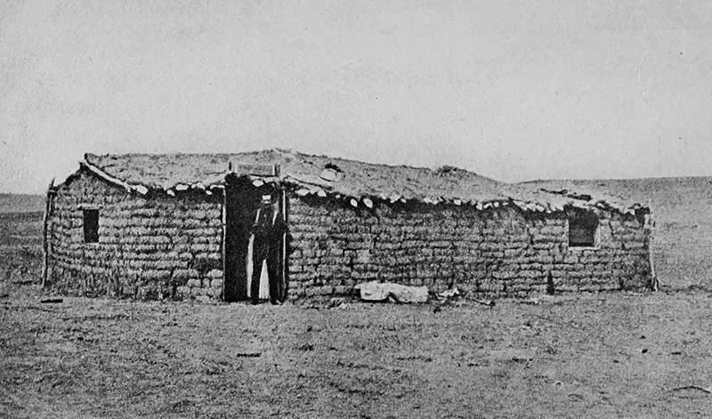 Black and white image of a sod house on a flat, barren landscape. A person stands in the doorway, wearing a hat and dark clothing. The house is made of stacked sod bricks with a rough roof. The setting appears desolate and historic.