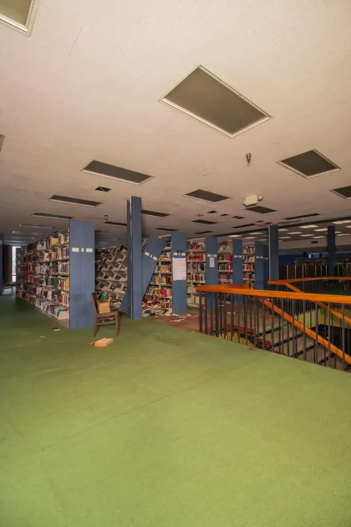 An interior of an empty library with green carpeted floors, wooden railings, and rows of bookshelves filled with books. Some books are scattered on the floor. The space is lit by overhead fluorescent lights.