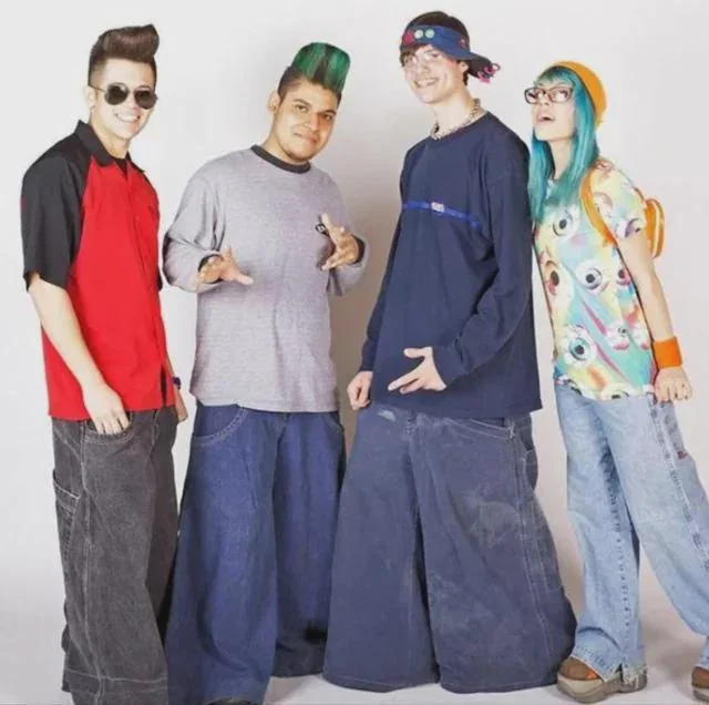 Four people stand together, each wearing oversized pants and colorful, eclectic tops. The first person sports sunglasses, the second has a tall mohawk, the third wears a beanie, and the fourth has turquoise hair and glasses. They all smile, posing casually.