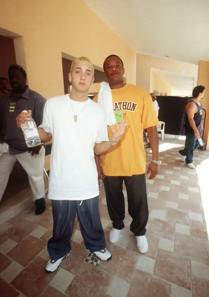 Two men pose in a hallway. The man on the left wears a white shirt, blue pants, and holds a water bottle. The man on the right wears an orange shirt and dark pants. Both display casual hand gestures. Other people are visible in the background.