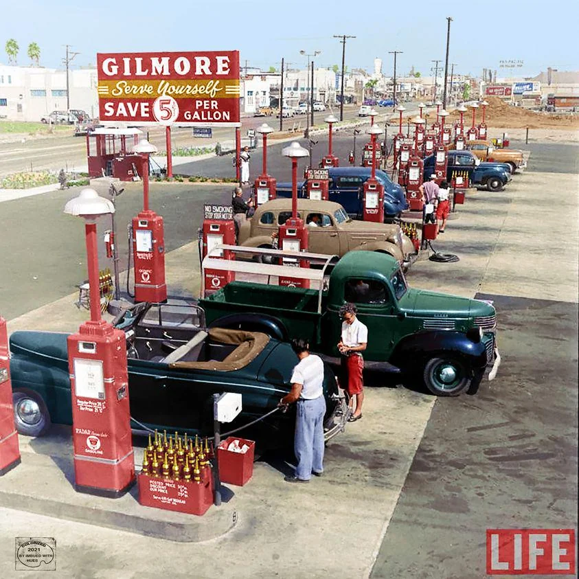 A vintage photograph of a Gilmore gas station with multiple cars lined up at red gas pumps. A woman and a man stand by a dark green convertible. The sign advertises gas for five cents off per gallon. The scene is lively with a retro vibe.