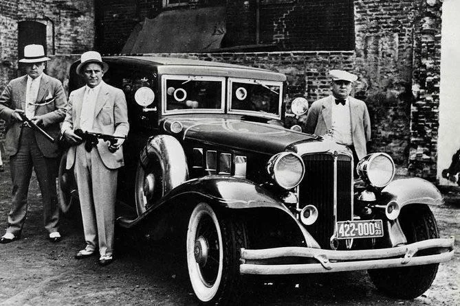 Three men in 1930s attire stand near a vintage car in an urban setting. Two men hold Tommy guns, wearing suits and hats. The car features a shiny exterior and a visible license plate with "422-0009". The background shows weathered brick walls.