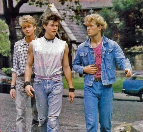 Three young men walk outdoors on a street. The first wears a plaid shirt, the second a sleeveless silver top, and the third a denim jacket with jeans. Trees and a stone building are in the background.