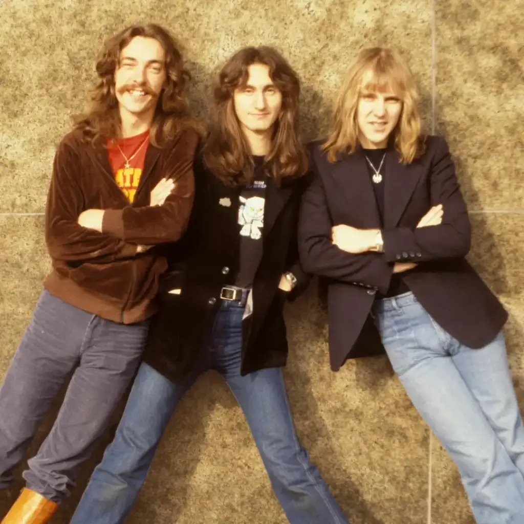 Three people with long hair stand in front of a textured wall. The one on the left wears a brown jacket, the middle wears a black jacket with a graphic tee, and the right wears a black blazer and blue jeans. All have their arms crossed and are smiling.