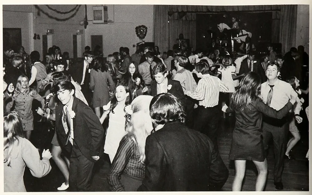 A black and white photo shows a crowded dance floor in a vintage setting. Men and women in 1970s attire are dancing energetically, with a band playing on a stage in the background. The room is decorated with streamers and a large wall mask.