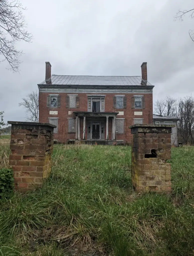 A dilapidated brick mansion stands with boarded windows and a metal roof, surrounded by overgrown grass. Two crumbling brick pillars are in the foreground, and the sky is overcast.