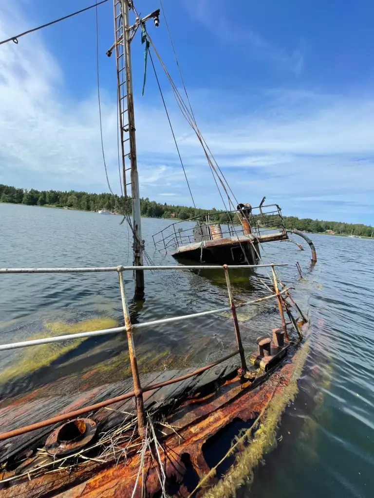 A rusted, partially submerged sailboat tilts to one side in a calm lake surrounded by trees. The mast is intact, but the deck is deteriorated, covered in moss and algae. The sky is clear with scattered clouds.