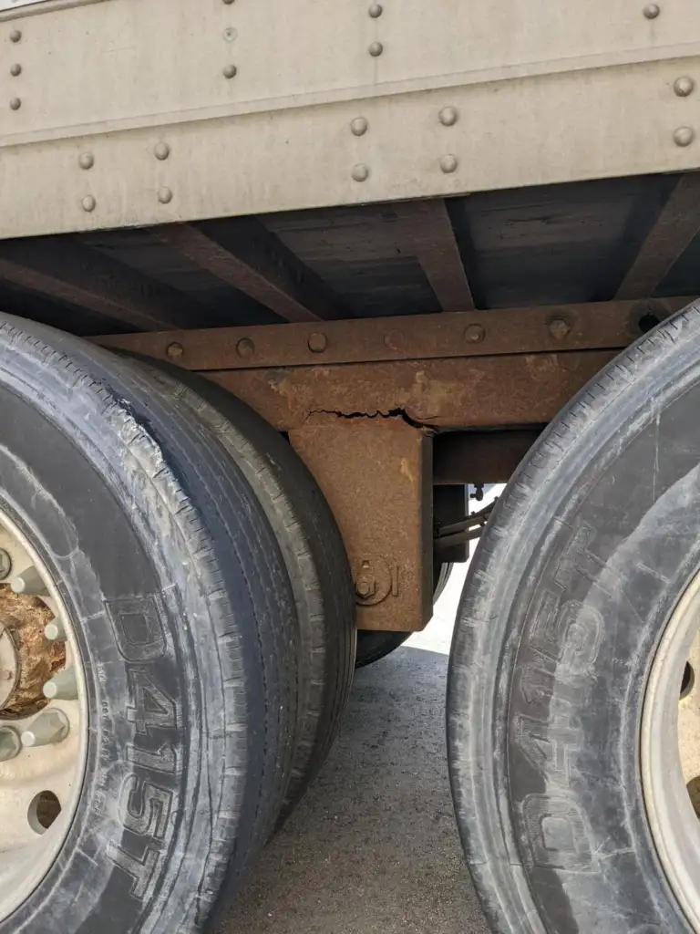 Close-up of a truck's rear wheels, showing signs of wear and surface rust on the metal components. The focus is on the suspension area, highlighting a crack on the rusty part above the tires.