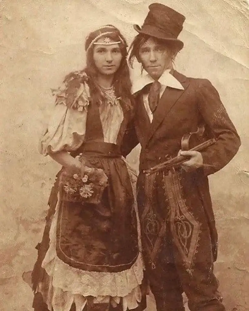 Vintage sepia photograph of two people standing together. The person on the left is wearing an elaborate dress with floral decorations and holding flowers. The person on the right is in a suit and top hat, holding a violin by their side.