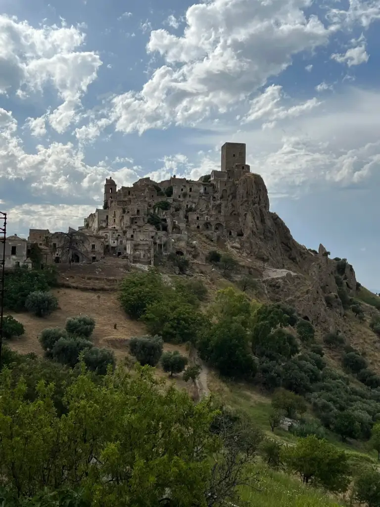 A hilltop town with stone buildings and a tower sits under a partly cloudy sky. The landscape surrounding the town features green trees and grassy areas, adding contrast to the rugged terrain.