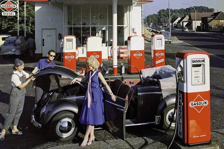 A vintage scene at a gas station shows a woman in a blue dress beside a classic convertible car with its hood open. Two attendants are assisting with the car. The station has red "Gasolin" pumps, and a building is visible in the background.