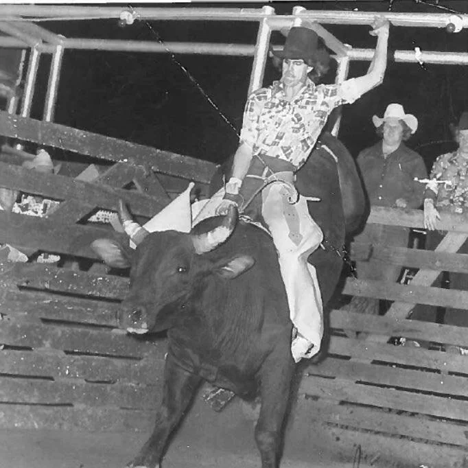 A black and white image of a rodeo scene with a person wearing a plaid shirt and hat riding a bull. The rider is in mid-action, holding on with one hand. Spectators in cowboy hats are visible in the background behind a wooden fence.