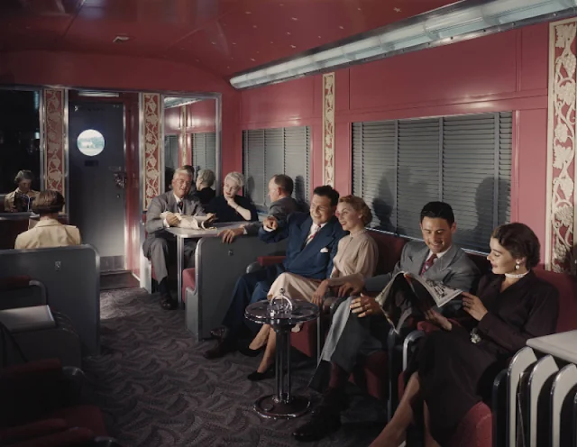 A vintage train car interior with elegantly dressed passengers. Men and women sit on upholstered seats, some chatting and laughing. The decor includes patterned walls and carpet. A round window on the door adds to the retro ambiance.