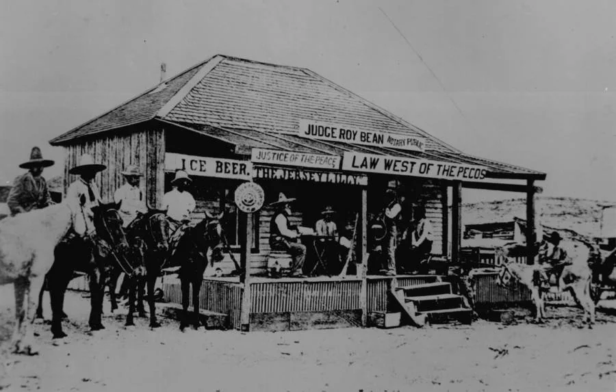 Black and white image of a rustic saloon labeled "Law West of the Pecos" with people seated on the porch. Several men on horseback stand nearby. Signs read "Judge Roy Bean" and "Ice Beer." The building appears weathered and old.