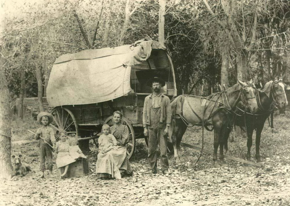 A black and white photo depicts a family from an earlier era, with a covered wagon and two horses. A man, woman, and children pose beside the wagon surrounded by trees. A dog sits near them on the leaf-covered ground.