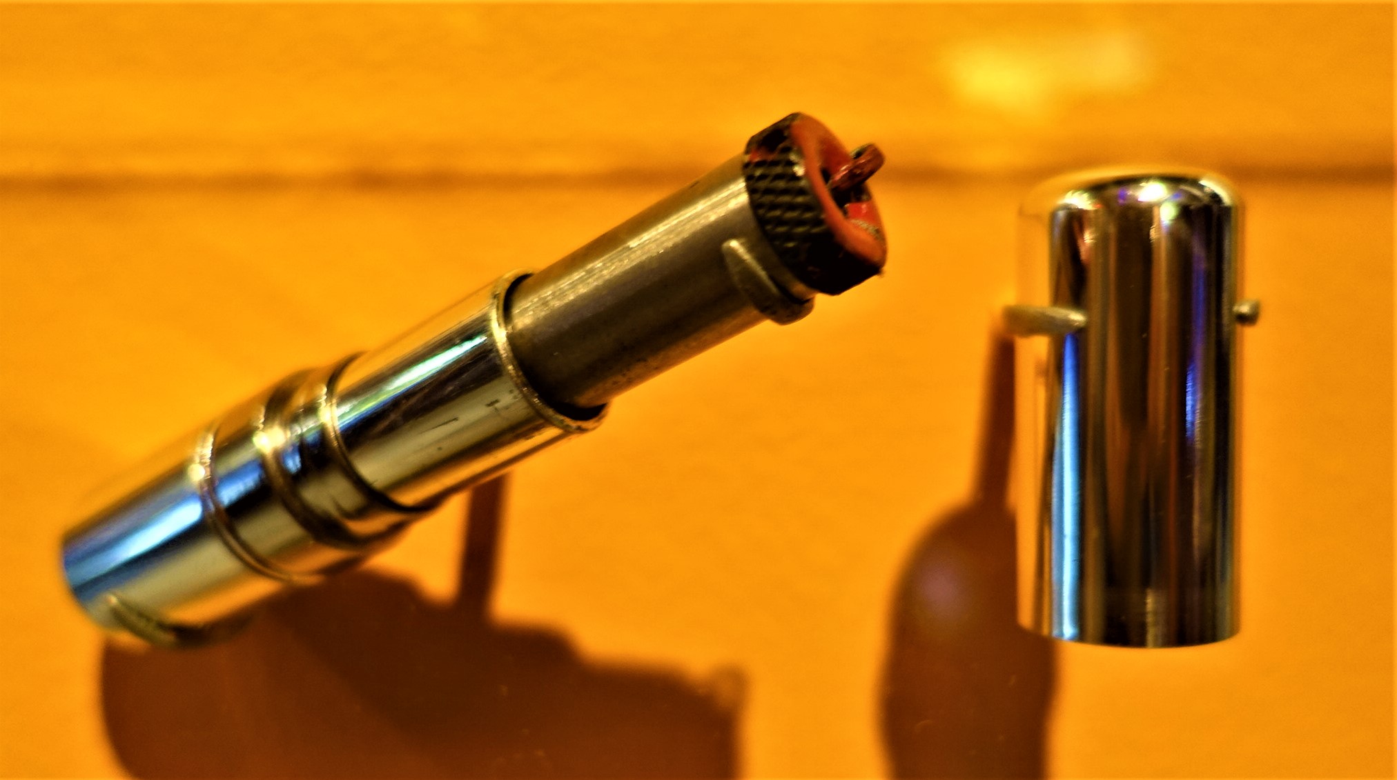 Close-up of a metallic cylinder device with an attached textured wheel on one end. The device is placed vertically against a warm yellow background. A separate, cylindrical cap with a small knob is nearby, casting a shadow.