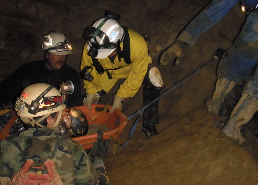 Rescue workers in helmets and headlamps carefully move a stretcher in a cave. The team navigates a narrow passage, using a blue rope for support. One person in a yellow jacket assists in steadying the stretcher.