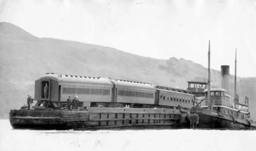 A black and white photo shows a train ferry transporting train cars across a body of water. The ferry has smokestacks and several people are visible on the deck. Mountains can be seen in the background under a cloudy sky.