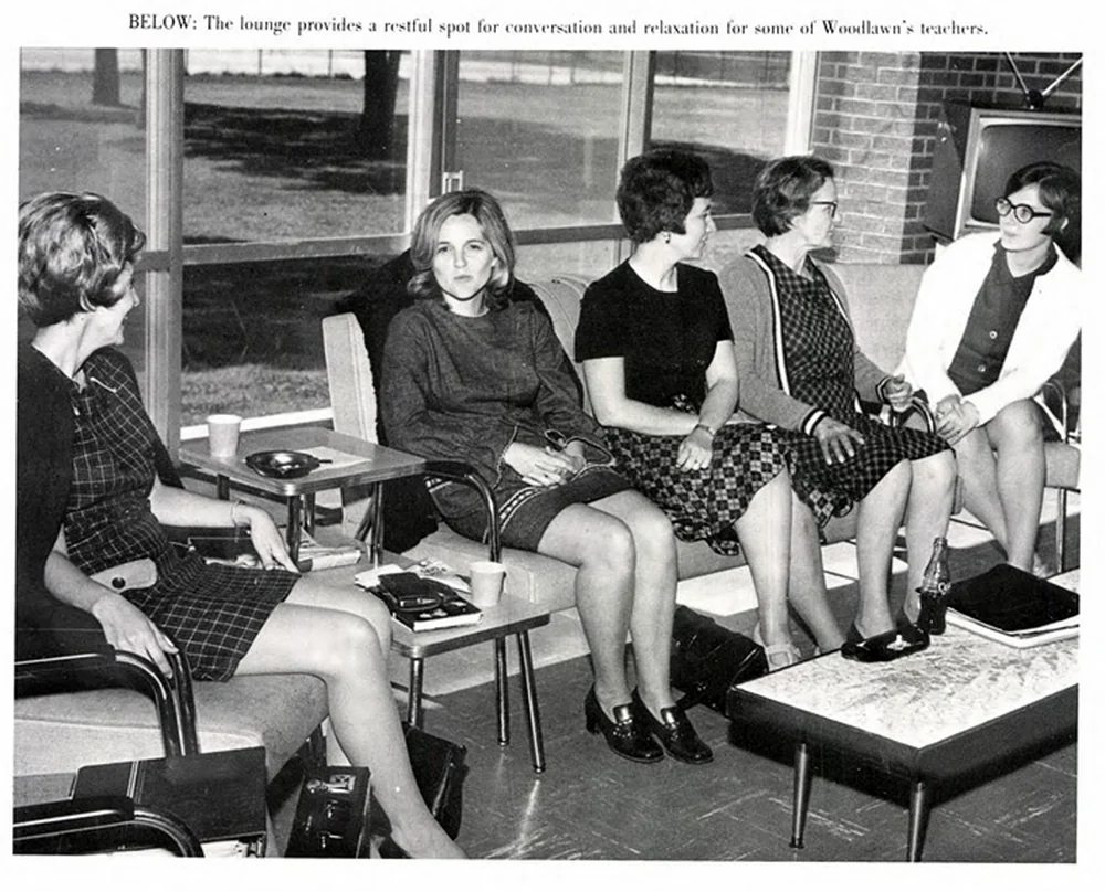 Five women seated in a lounge, engaged in conversation. The room has large windows with a view outside. They are casually dressed, and two have crossed legs. There are coffee cups on small tables in front of them.