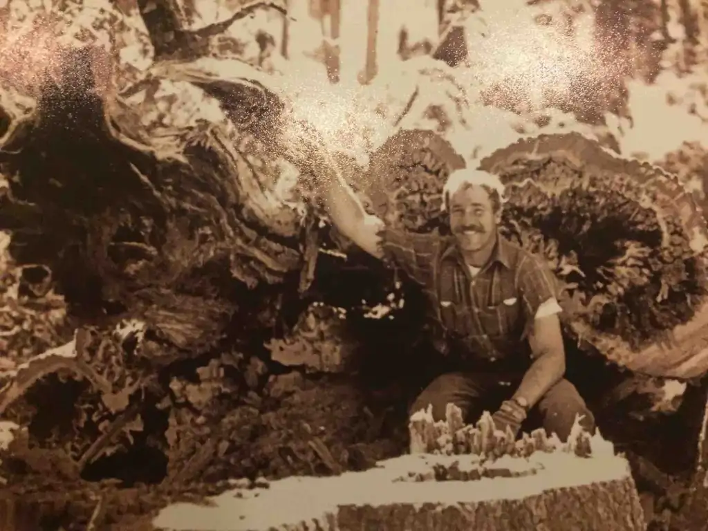 A man with a mustache, wearing a plaid shirt and pants, squats next to a large fallen tree trunk in a forest. He rests one arm on the tree, surrounded by nature. The image is in sepia tone.